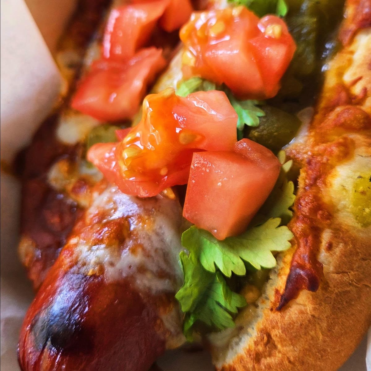 A DogCraft hot dog in a toasted bun under melted cheese, diced tomato and fresh cilantro.